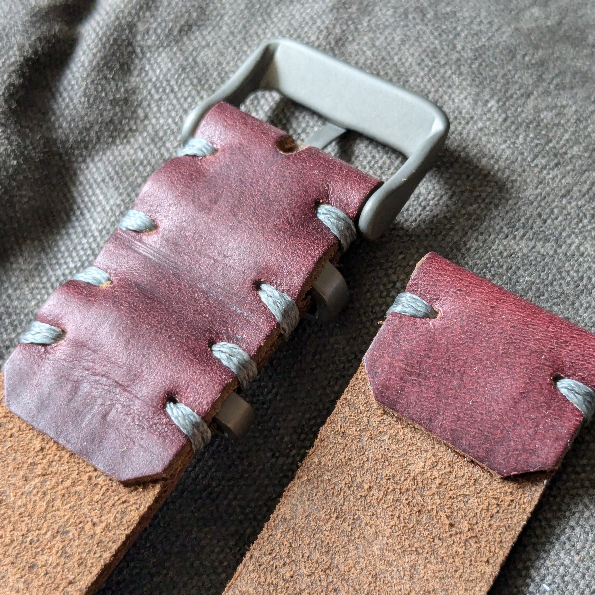 A close-up of a handmade Horween Color 8 leather watch strap, light brown leather strap, and a titanium buckle, laid on a textured gray fabric background.