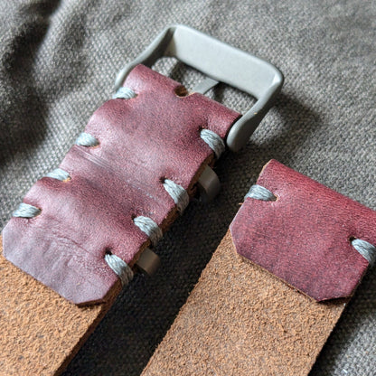 A close-up of a handmade Horween Color 8 leather watch strap, light brown leather strap, and a titanium buckle, laid on a textured gray fabric background.