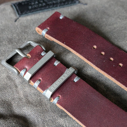 A close-up of a Horween Color 8 leather watch strap with a silver-toned metal buckle and hardware, laid on a textured gray fabric background.